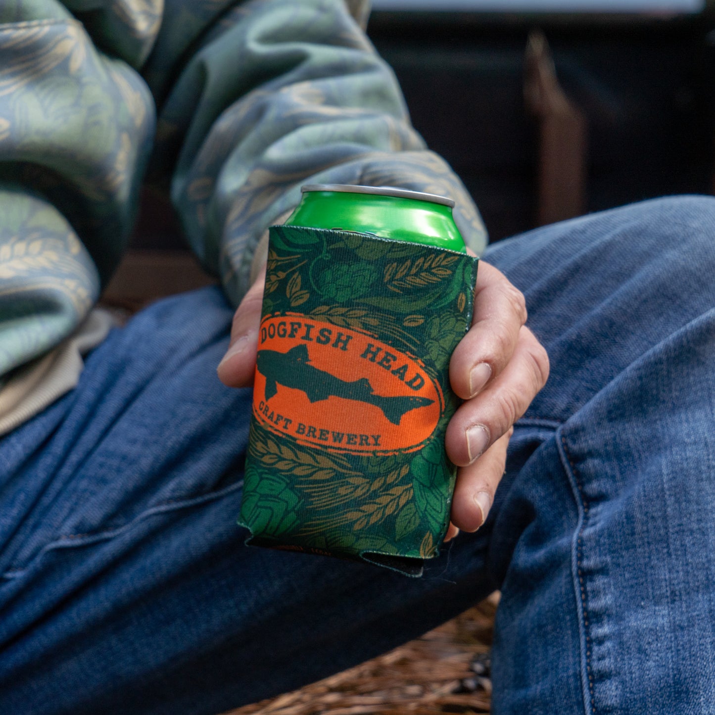Being held with a can in it, is a green camo can cooler featuring an orange Dogfish Head shark and shield logo.