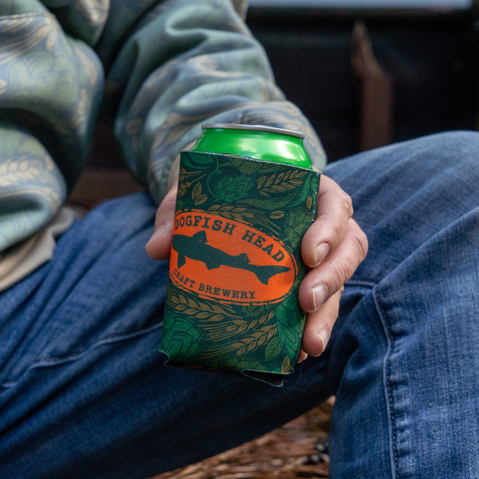 Being held with a can in it, is a green camo can cooler featuring an orange Dogfish Head shark and shield logo.