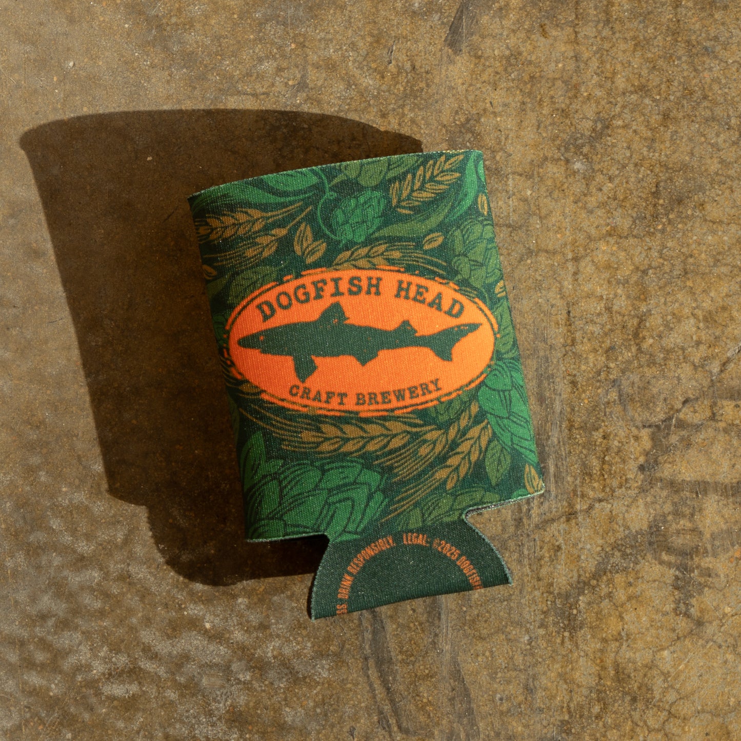 Laid flat on a surface, is a green camo can cooler featuring an orange Dogfish Head shark and shield logo.