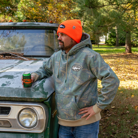 A person wearing a green camo hoodie featuring a black Dogfish Head shark and shield logo on the left side of the chest. Person is wearing an orange Dogfish beanie and is holding the matching camo can cooler.