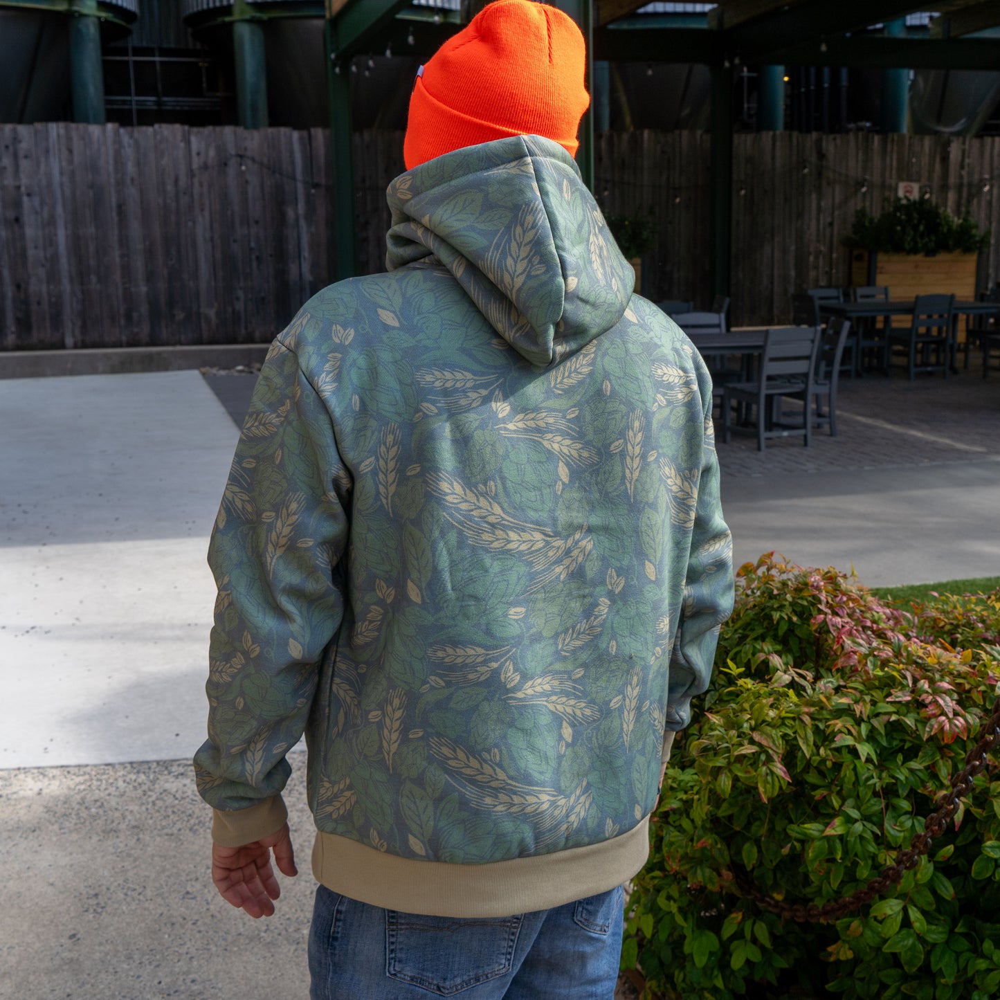 A person wearing a green camo hoodie modeling to show the back. Person is wearing an orange Dogfish beanie and is holding the matching camo can cooler.