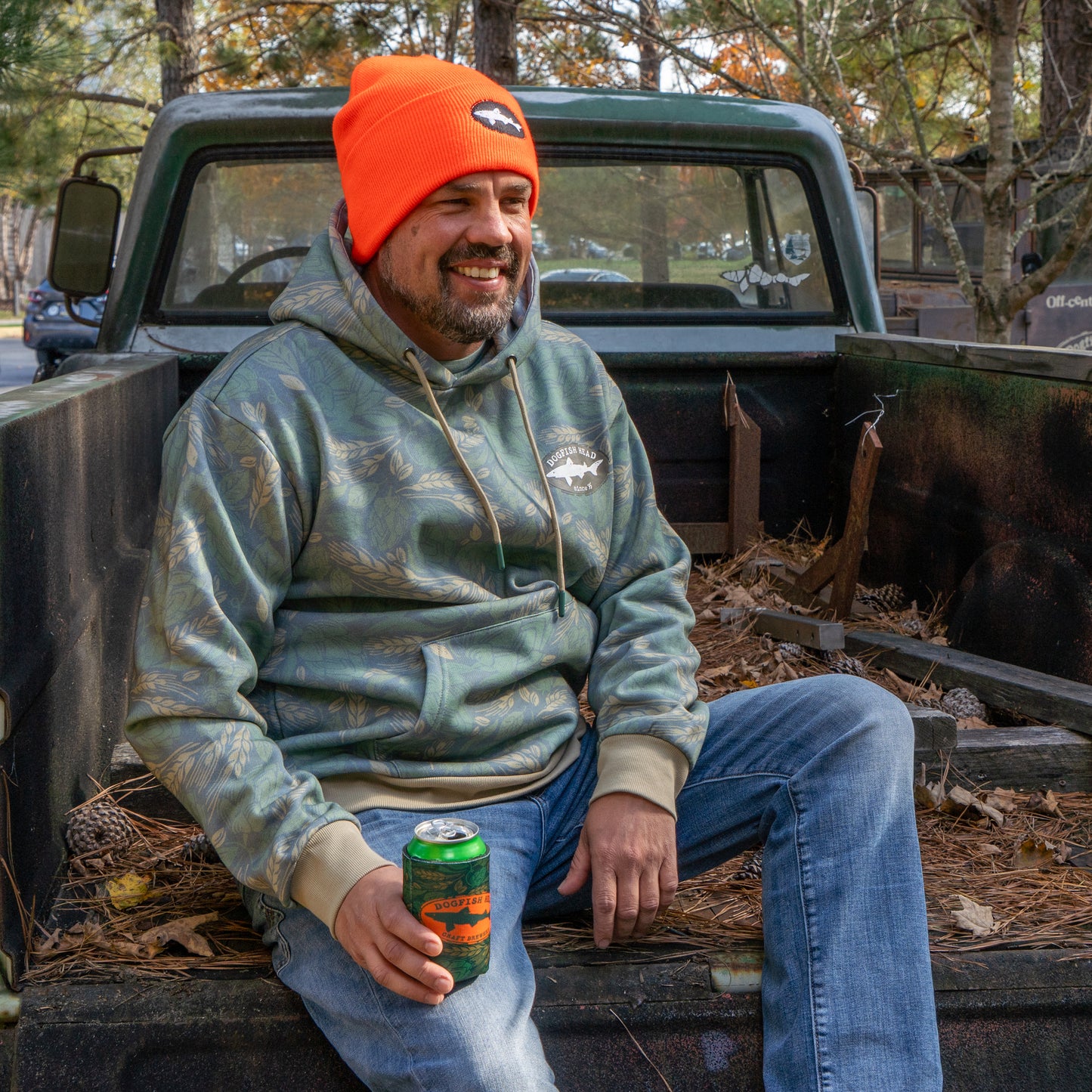 A person wearing a green camo hoodie featuring a black Dogfish Head shark and shield logo on the left side of the chest. Person is wearing an orange Dogfish beanie and is holding the matching camo can cooler.
