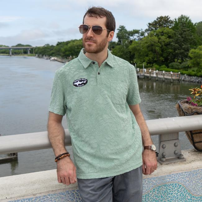 A man in sunglasses and a Howler Brothers Mint Terry Polo leans against a bridge railing overlooking a river, with trees and greenery in the background.