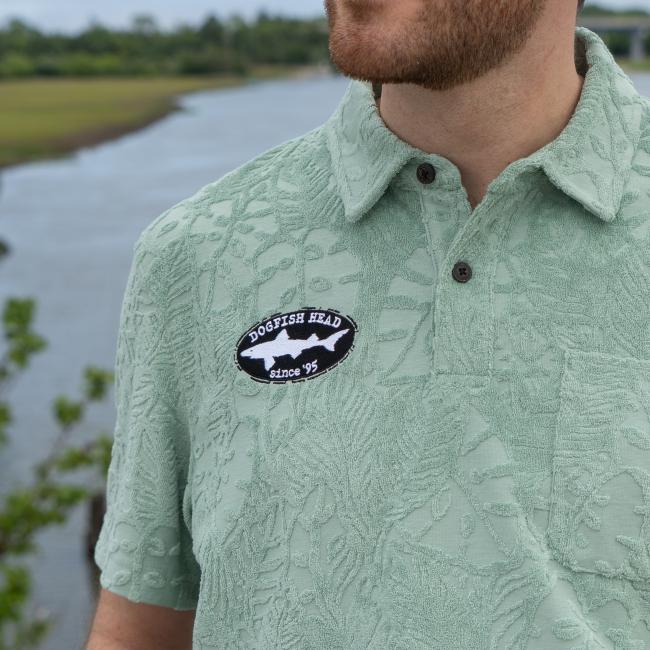 A man stands outdoors near water and greenery, wearing a Howler Brothers Mint Terry Polo with a Dogfish Head since 95 patch. Only his torso and part of his face are visible.