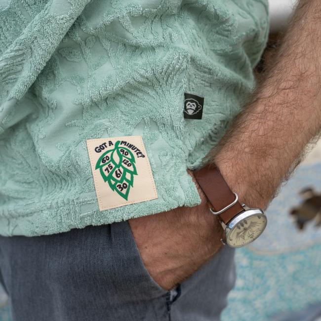 A person wears the Howler Brothers Mint Terry Polo—a light green textured shirt with a "Got a Minute?" patch and green clock design—paired with blue jeans, a brown leather watch, and their hand in their pocket.