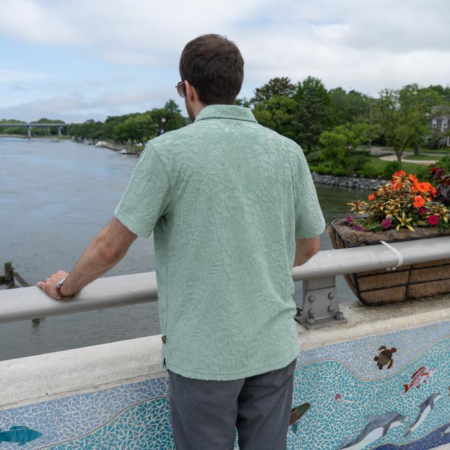 A man in a Howler Brothers Mint Terry Polo and gray pants stands on a bridge, framed by mosaic tiles, blooming planters, and trees as clouds gather—a scene perfect for Howler Brothers style.