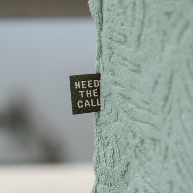 A close-up of the Howler Brothers Mint Terry Polo shows its textured light green fabric and a small black "HEED THE CALL" tag, while the blurred background reflects the adventurous spirit of Dogfish Head.