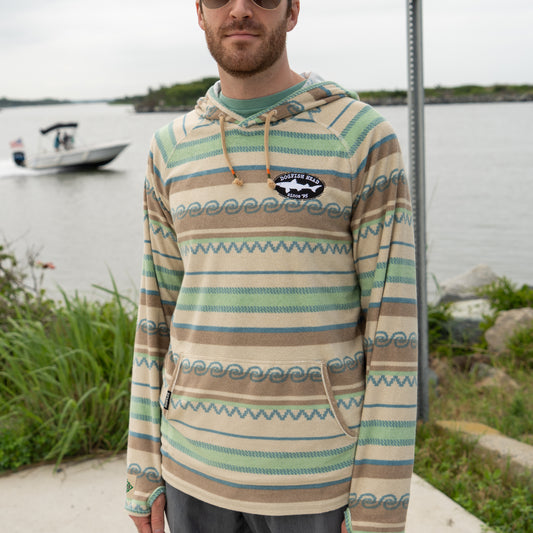 A man in sunglasses and a Howler Brothers Striped Terry Cloth Hoodie stands by the water, with a boat in the background and greenery surrounding him.