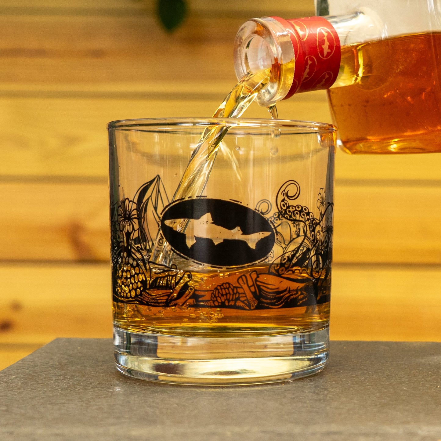 A Hull and Helm Rocks Glass by Grandstand Glassware & Apparel, featuring a fish logo and black designs, is filled with amber whiskey against a rustic wooden background.