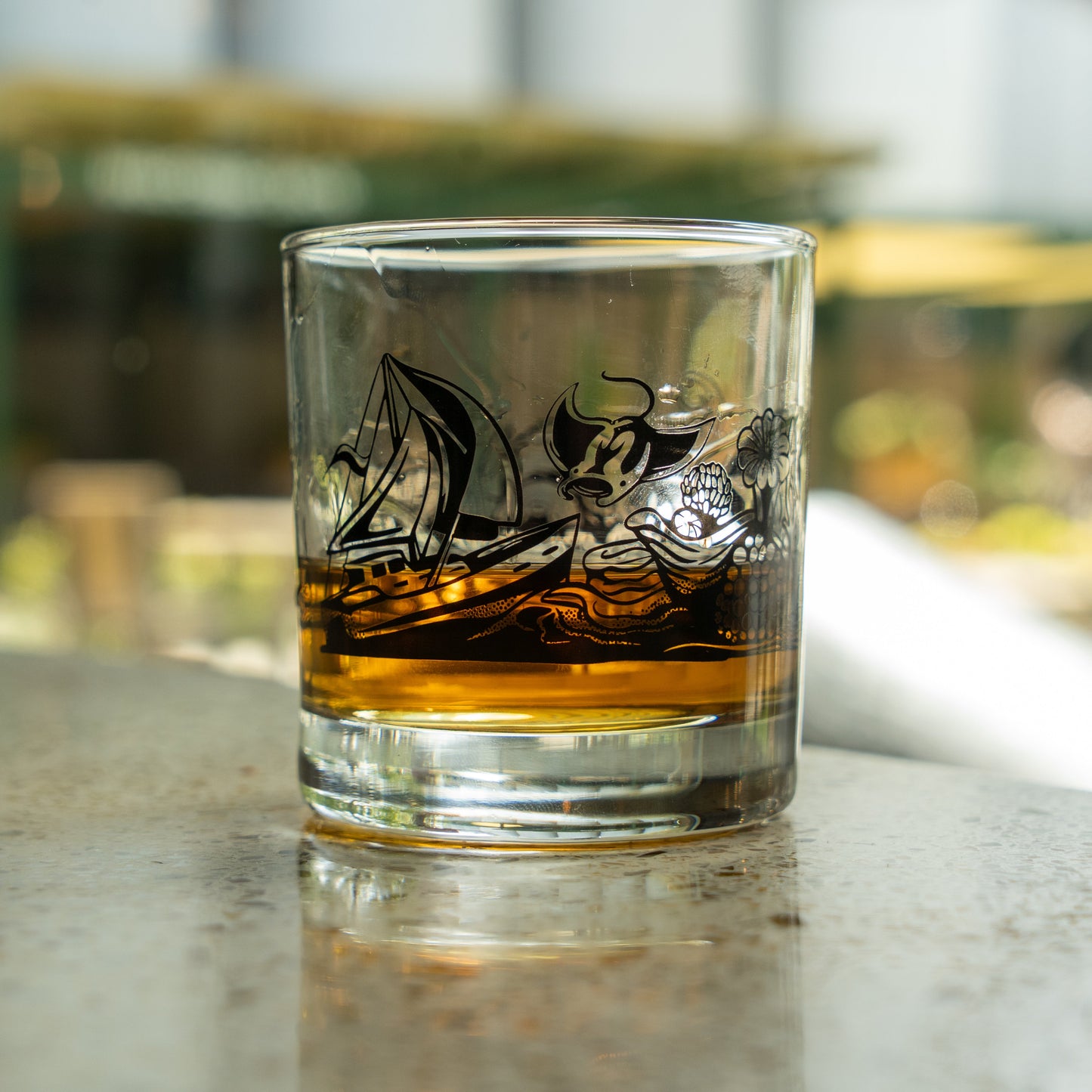 A Hull and Helm Rocks Glass by Grandstand Glassware & Apparel, featuring black nautical designs with a sailboat and waves, holds amber whiskey on a reflective surface against a sunlit, blurred background.