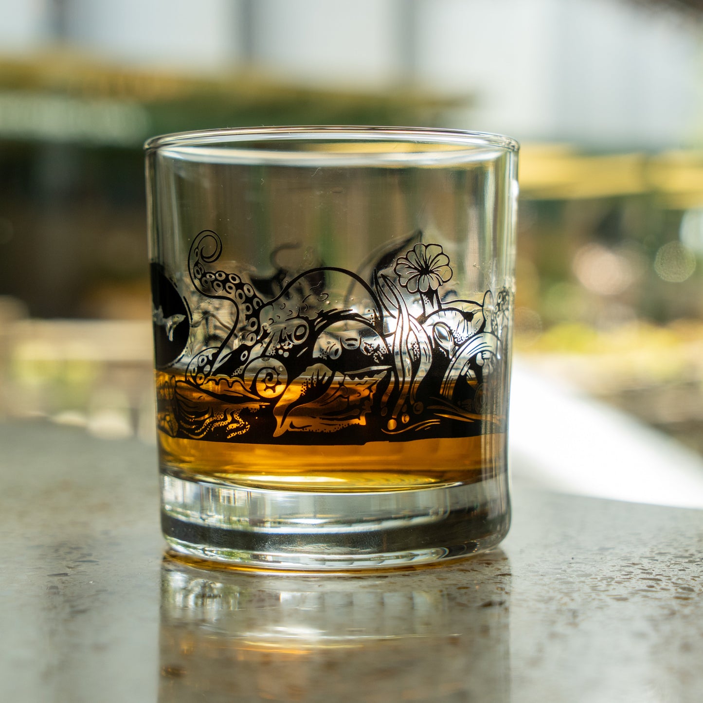 The Hull and Helm Rocks Glass by Grandstand Glassware & Apparel rests on a reflective surface, holding amber whiskey. Black octopus tentacle and sea plant illustrations decorate the glass; the background is softly blurred.