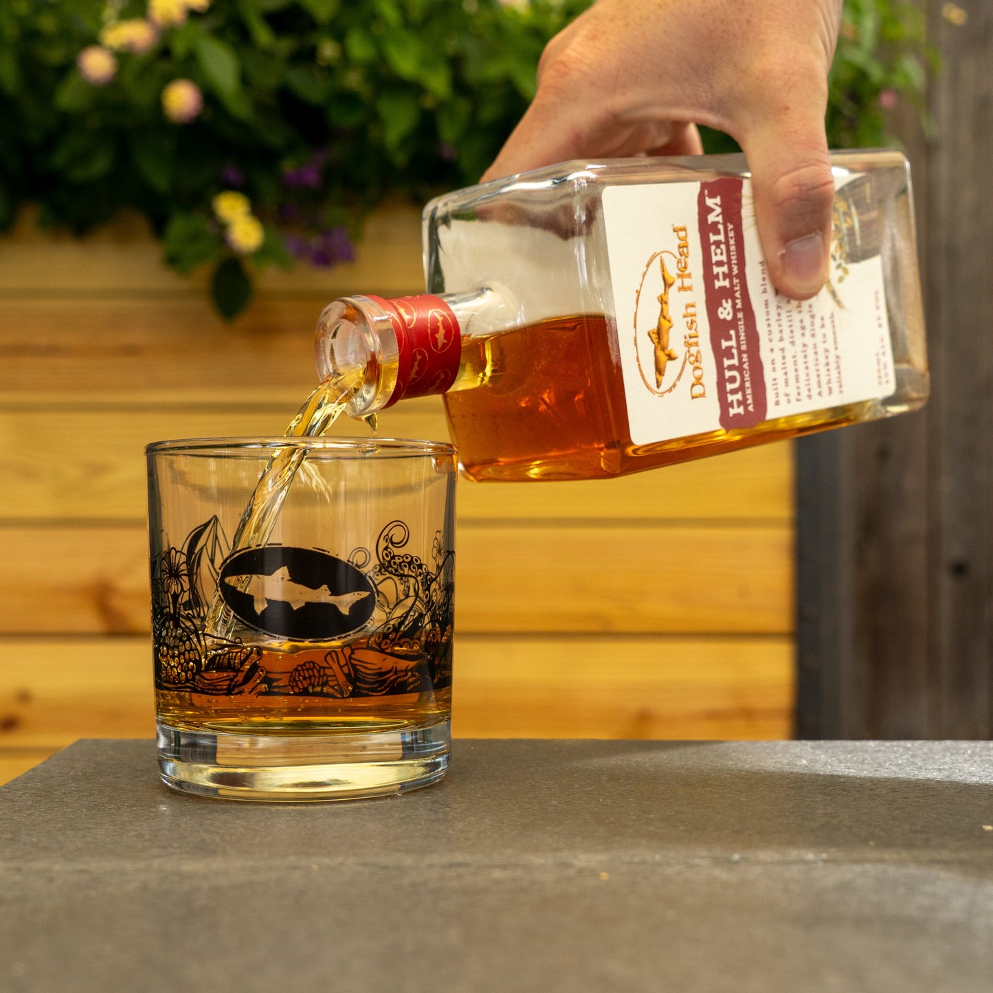 A hand pours whiskey into a Hull and Helm Rocks Glass by Grandstand Glassware & Apparel, featuring a fish logo, set outdoors with a wooden fence and flowers, highlighting the glass’s quality craftsmanship.