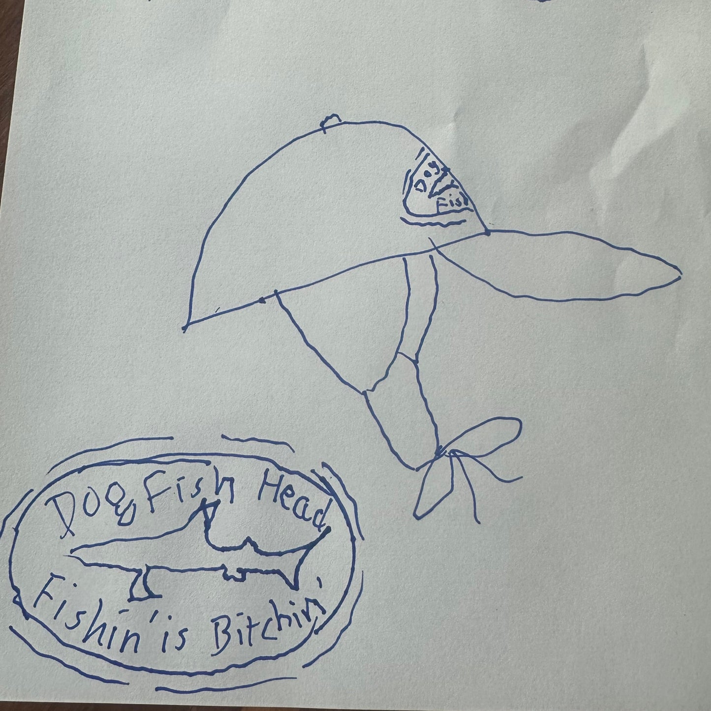 A hand-drawn blue pen illustration of the Fishin' is Bitchin' Retro Hat with Ear Flaps by Grandstand Glassware & Apparel, featuring a fish and the word "Fish" on the crown. Beneath, the text "Dogfish Head Fishin’ Is Bitchin’" appears alongside an oval design with a shark, reminiscent of the iconic Dogfish Head flap hat.