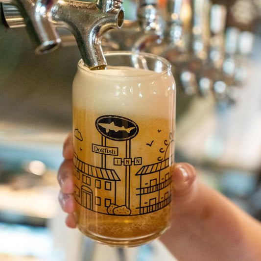 A hand holds the INN Can Glass by Grandstand Glassware & Apparel as beer pours in, forming a foamy head. The glass features a whimsical design of buildings and a Dogfish Head INN street sign celebrating Lewes, Delaware.