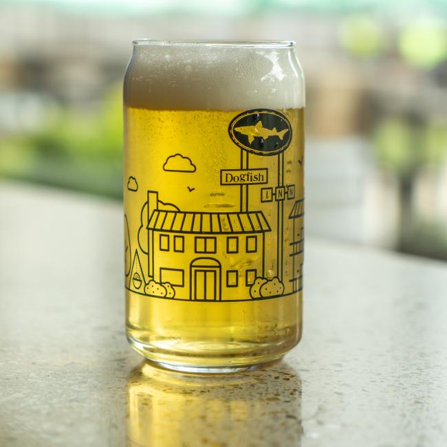 A Grandstand Glassware & Apparel INN Can Glass, filled with light amber beer and topped with foam, sits on a countertop. The glass displays a black line illustration of Lewes, Delaware with the Dogfish Head INN. The background appears blurred.