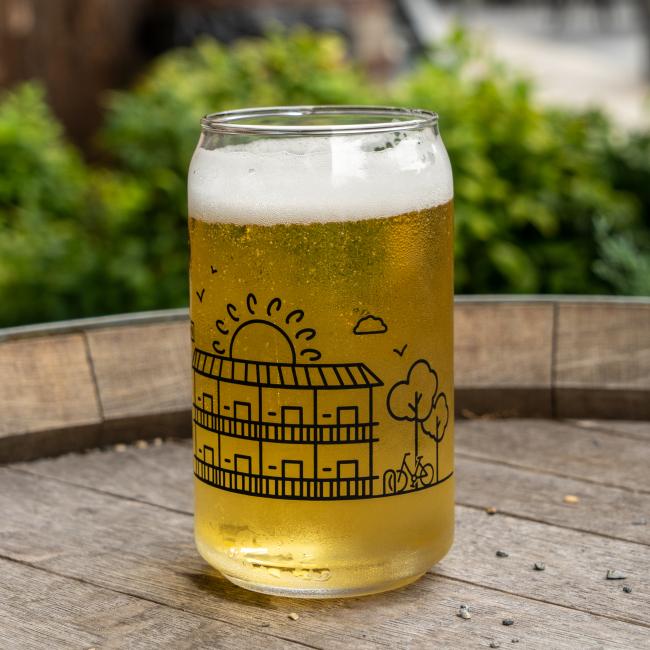 A clear INN Can Glass by Grandstand Glassware & Apparel, filled with light amber beer, rests on a wooden surface outdoors at Dogfish Head INN in Lewes, DE. The glass displays a minimalist line drawing of a building, sun, and trees against blurred green foliage.
