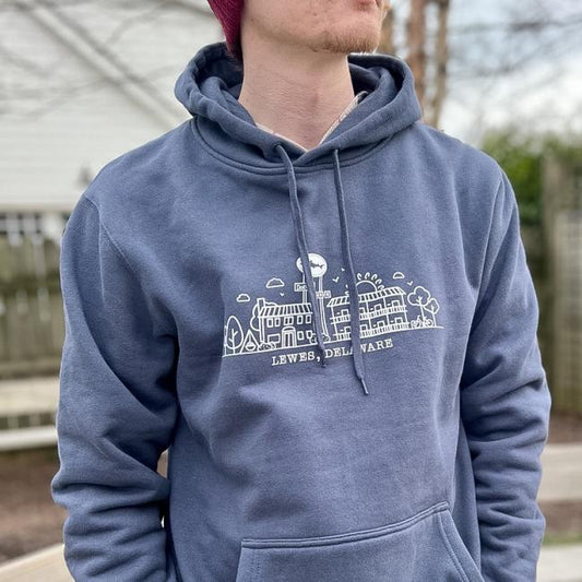 A person in a maroon beanie and the GO WEST T-SHIRT COMPANY INN Logo Blue Hoodie, which features a white line drawing of buildings and trees labeled LEWES, DELAWARE, stands outdoors with a blurred fence and trees in the background.