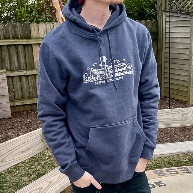 Outdoors in a fenced yard, someone wears the GO WEST T-SHIRT COMPANY's INN Logo Blue Hoodie featuring a white line drawing of buildings and "Lewes, Delaware" on the front—a perfect souvenir sweatshirt moment.