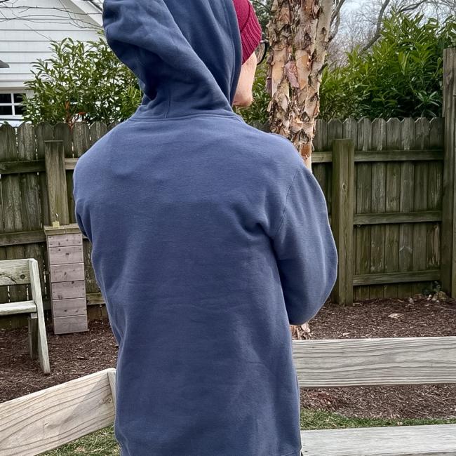 A person in the GO WEST T-SHIRT COMPANY INN Logo Blue Hoodie and a red beanie stands outside with their back to the camera, near a wooden fence, trees, and benches in a yard.