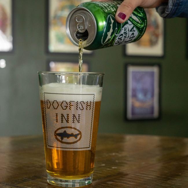 A hand pours beer into a Grandstand Glassware & Apparel INN Pint Glass labeled Dogfish INN, with framed artwork on the green wall at the cozy Dogfish INN in Lewes, Delaware.