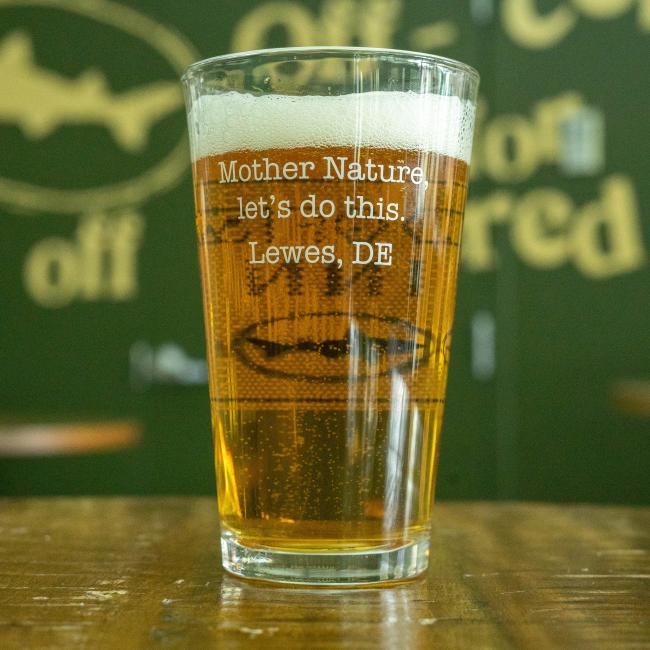 The INN Pint Glass by Grandstand Glassware & Apparel is filled with amber beer, featuring "Mother Nature, let’s do this. Lewes Delaware" text. A foamy top crowns the glass, set against a green background with blurred yellow lettering.