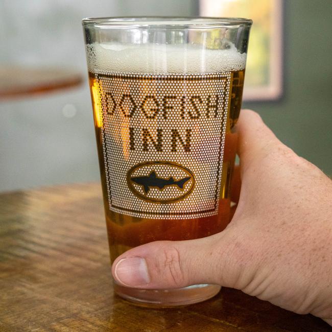 A hand holds the INN Pint Glass by Grandstand Glassware & Apparel, filled with beer and resting on a wooden table. The blurred background evokes the laid-back atmosphere of Lewes, Delaware.