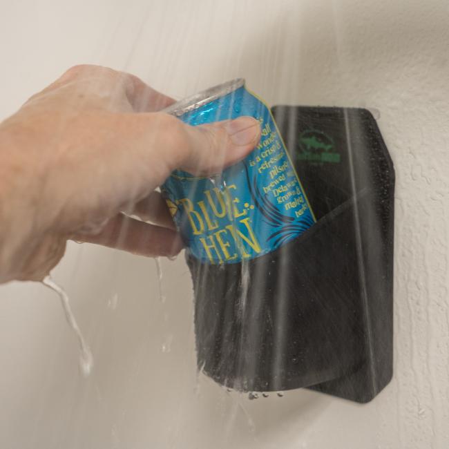A hand places a can of Blue Hen beer into a sleek black INN Shower Beer Holder by Sudski, mounted on the shower wall with water spraying nearby.