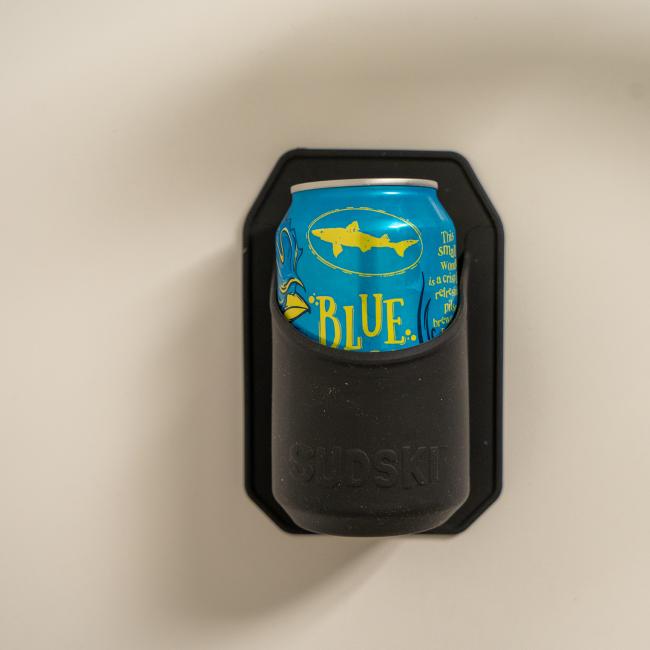 A blue beer can with yellow writing rests in a black wall-mounted INN Shower Beer Holder by Sudski, shown against a light beige background—an ideal gift for any beer lover.