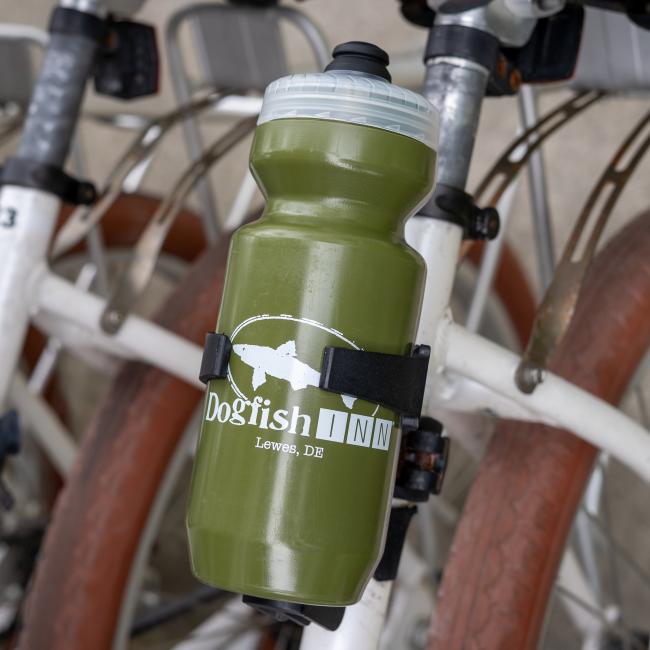 A green Specialized INN Water Bottle, BPA free, is attached to the frame of a white bicycle, with other bikes and brown tires in the background.