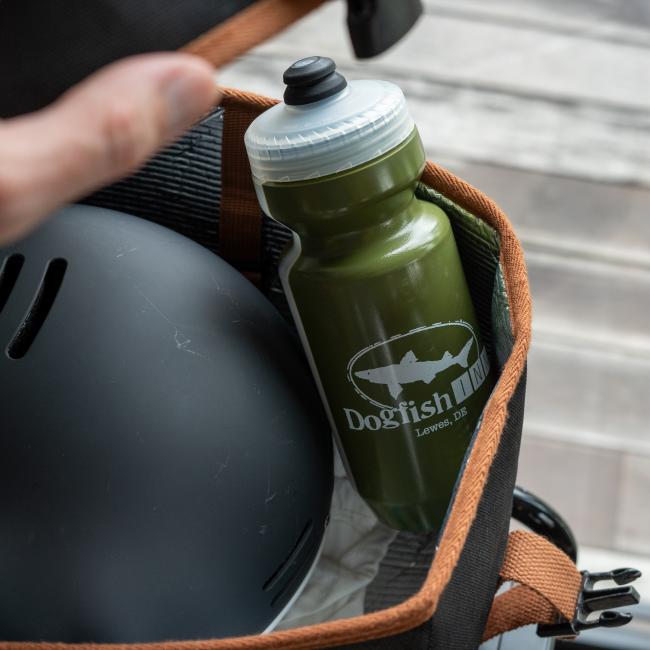 A hand holds open a brown bag on a wooden surface, revealing a black helmet and a green Specialized INN Water Bottle.