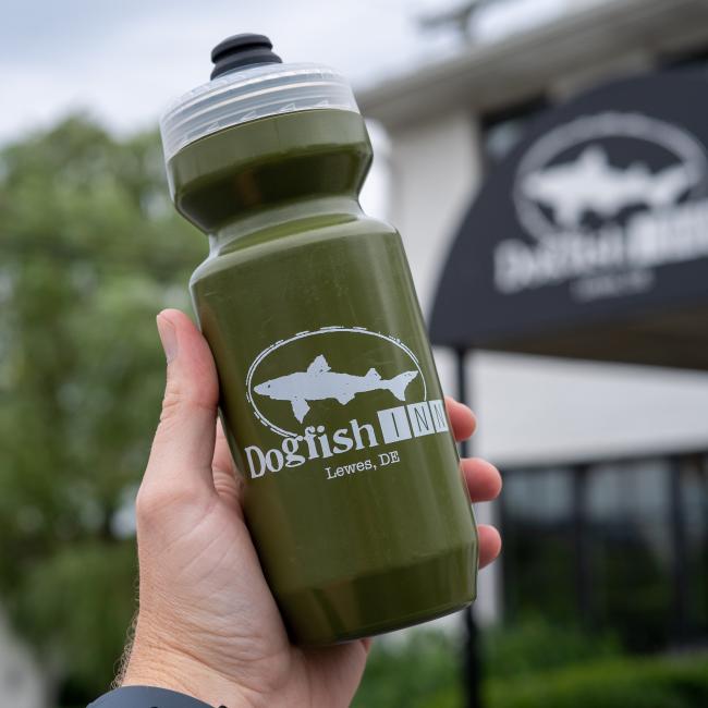 A hand holds a green INN Water Bottle by Specialized, featuring a white shark logo and "Dogfish Head INN, Lewes, DE." In the background, a building and matching sign display the same iconic logo.