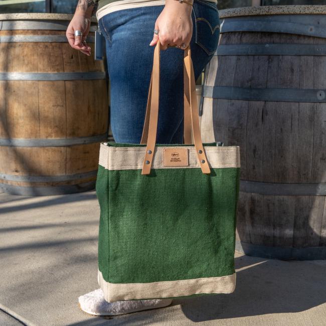 Green bag with a thick off-white border on the top and bottom of the bag. Straps are tan leather, and the top border features a small square leather patch with the Dogfish Head shark and shield logo and writing "Explore Goodness".