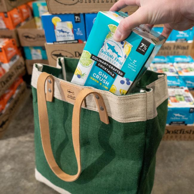 Green bag with a thick off-white border on the top and bottom of the bag. Straps are tan leather, and the top border features a small square leather patch with the Dogfish Head shark and shield logo and writing "Explore Goodness".