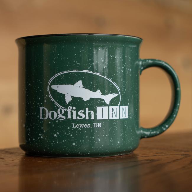 A dark green 12oz Inn Camp Cup by Grandstand Glassware & Apparel, featuring the Dogfish Inn logo with a fish silhouette and “Dogfish INN, Lewes, DE,” rests on a wooden surface.