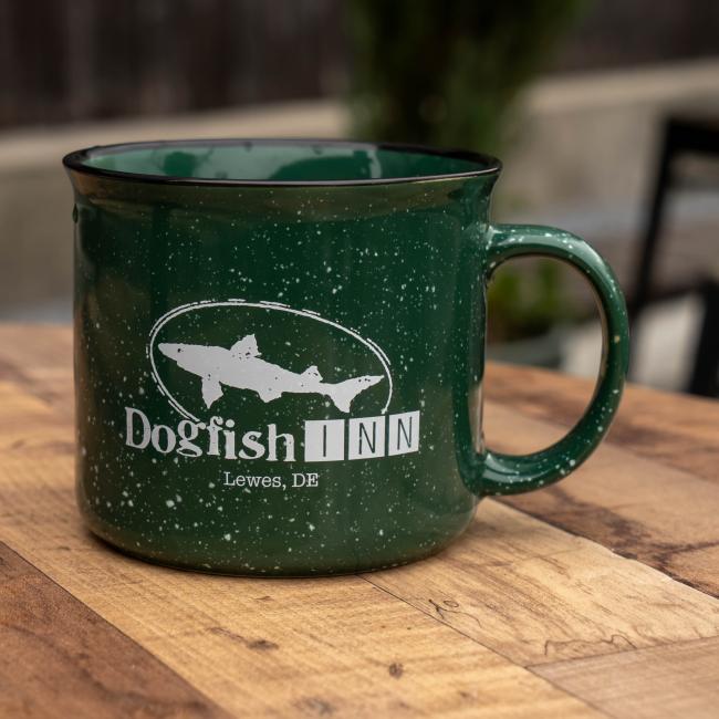 A dark green, 12oz Inn Camp Cup by Grandstand Glassware & Apparel with a handle rests on a wooden surface. The mug displays the white Dogfish Inn logo, featuring a fish graphic and "Dogfish INN Lewes, DE" text.