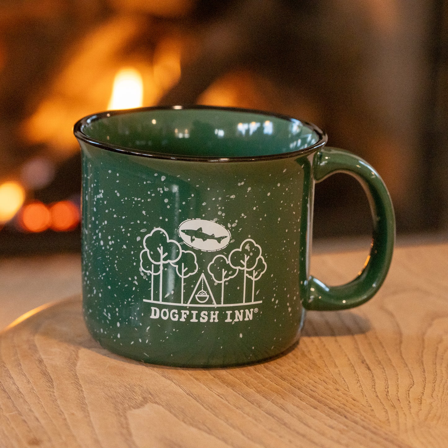 A green speckled 12oz Inn Camp Cup by Grandstand Glassware & Apparel, featuring the Dogfish Inn logo with trees and a fish, rests on a wooden surface lit by the warm glow of a blurred fire.