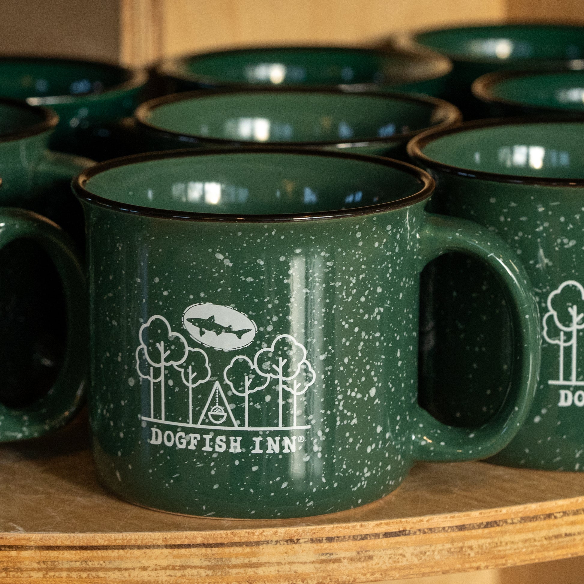 Several green speckled 12oz Inn Camp Cups by Grandstand Glassware & Apparel, each featuring "Dogfish Inn" and a white outdoor logo, are stacked close together on a wooden shelf.