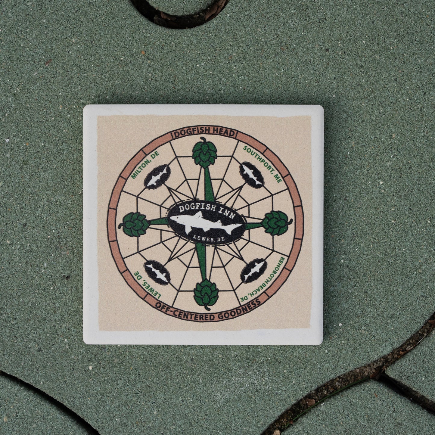 The Inn Ceramic Off Centered Coaster by Paint the Town, Inc. features hops, fish, and compass-style art with "Dogfish Inn Lewes, DE"—perfect for craft lovers—resting stylishly on a green tiled surface.