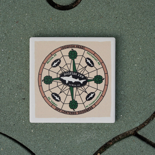 The Inn Ceramic Off Centered Coaster by Paint the Town, Inc. features hops, fish, and compass-style art with "Dogfish Inn Lewes, DE"—perfect for craft lovers—resting stylishly on a green tiled surface.