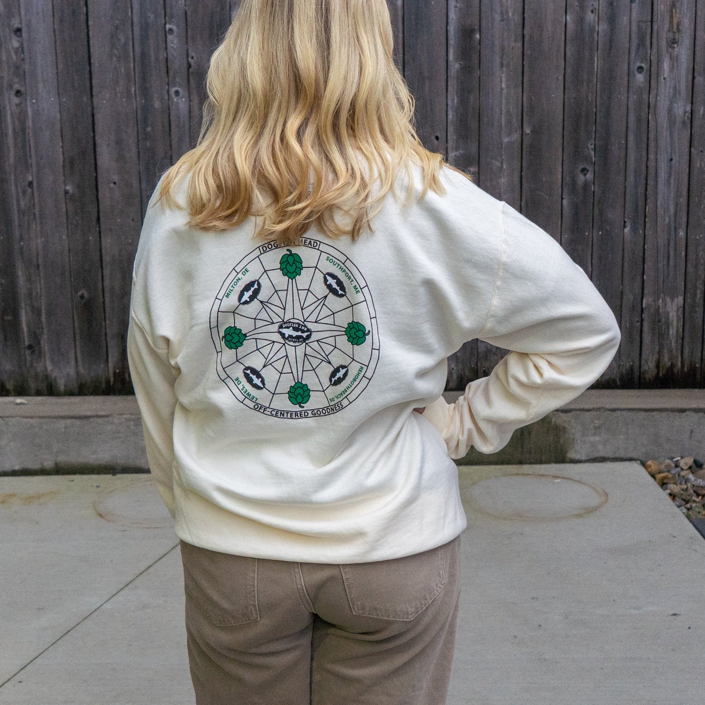 A blonde person in brown pants stands outdoors facing a wooden fence, wearing the Inn Cream Compass Crew by GO WEST T-SHIRT COMPANY—a white ring-spun cotton hoodie with a circular back graphic.