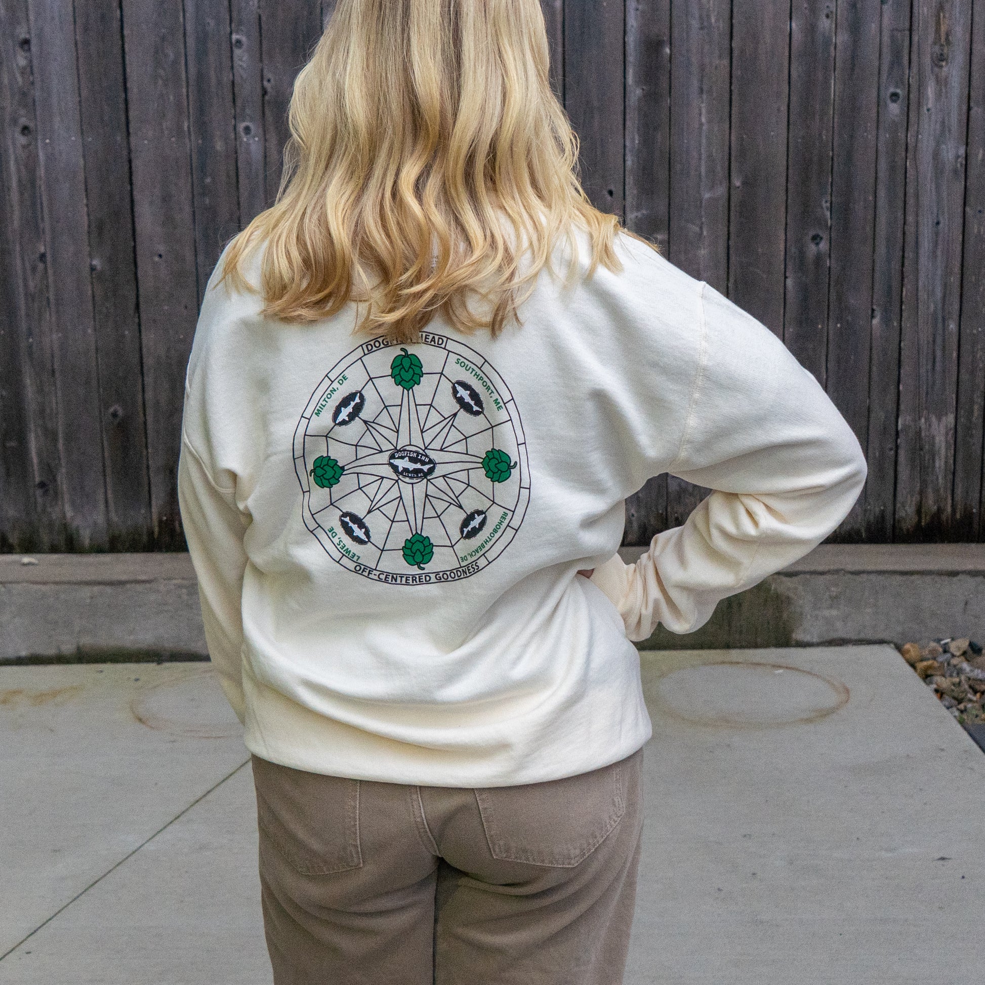 A blonde person in brown pants stands outdoors facing a wooden fence, wearing the Inn Cream Compass Crew by GO WEST T-SHIRT COMPANY—a white ring-spun cotton hoodie with a circular back graphic.