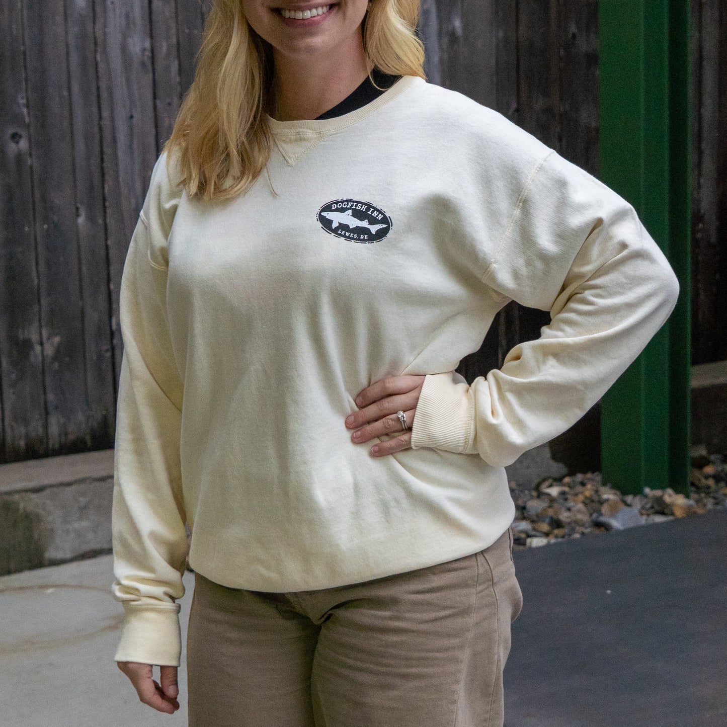 A person with long blonde hair, wearing the GO WEST T-SHIRT COMPANY Inn Cream Compass Crew and khaki pants, stands smiling with one hand on their hip in front of a wooden fence.