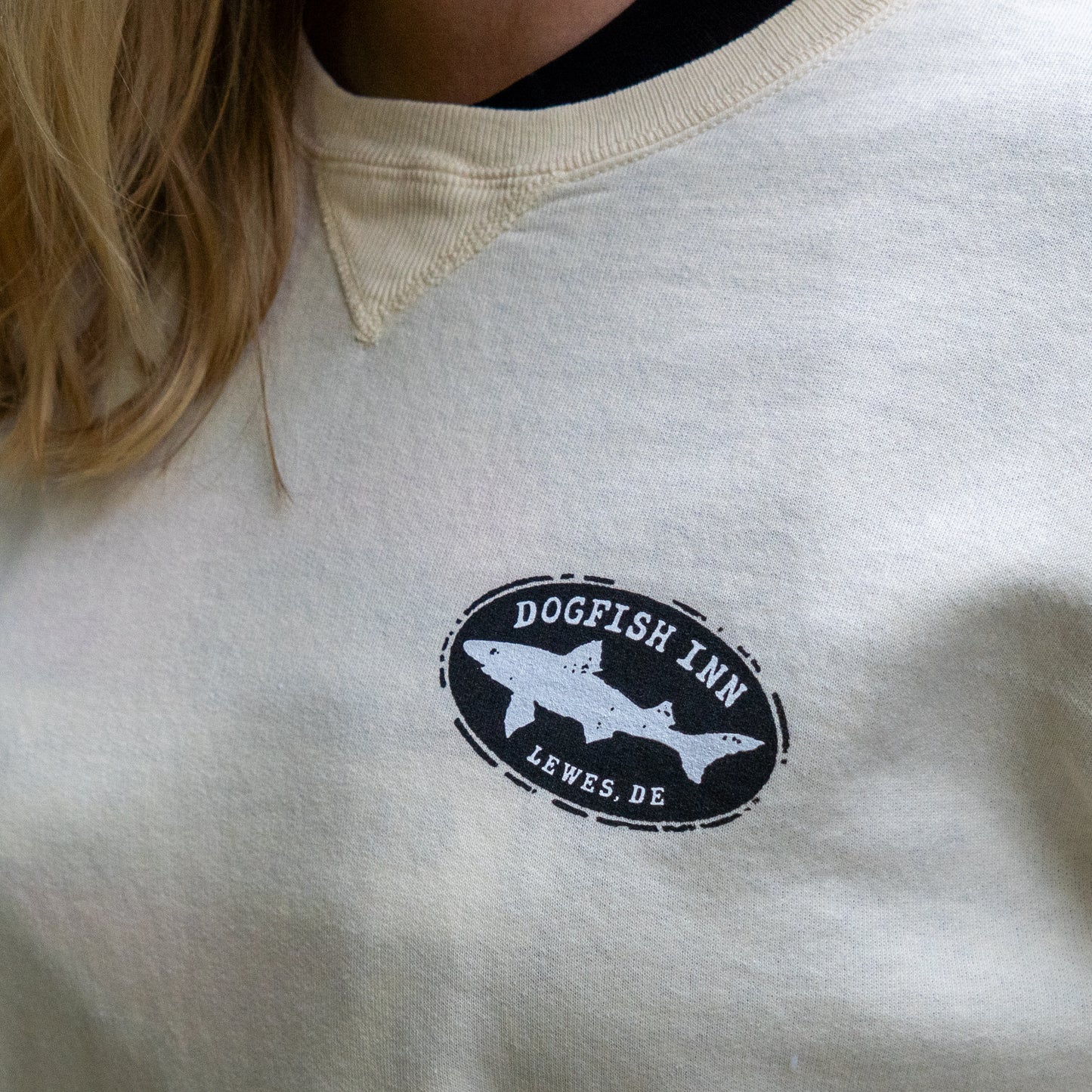 Close-up of a person wearing the Inn Cream Compass Crew by GO WEST T-SHIRT COMPANY, featuring a beer compass logo with a fish and "Dogfish Inn Lewes, DE" on the left chest. Part of their long blonde hair is visible.