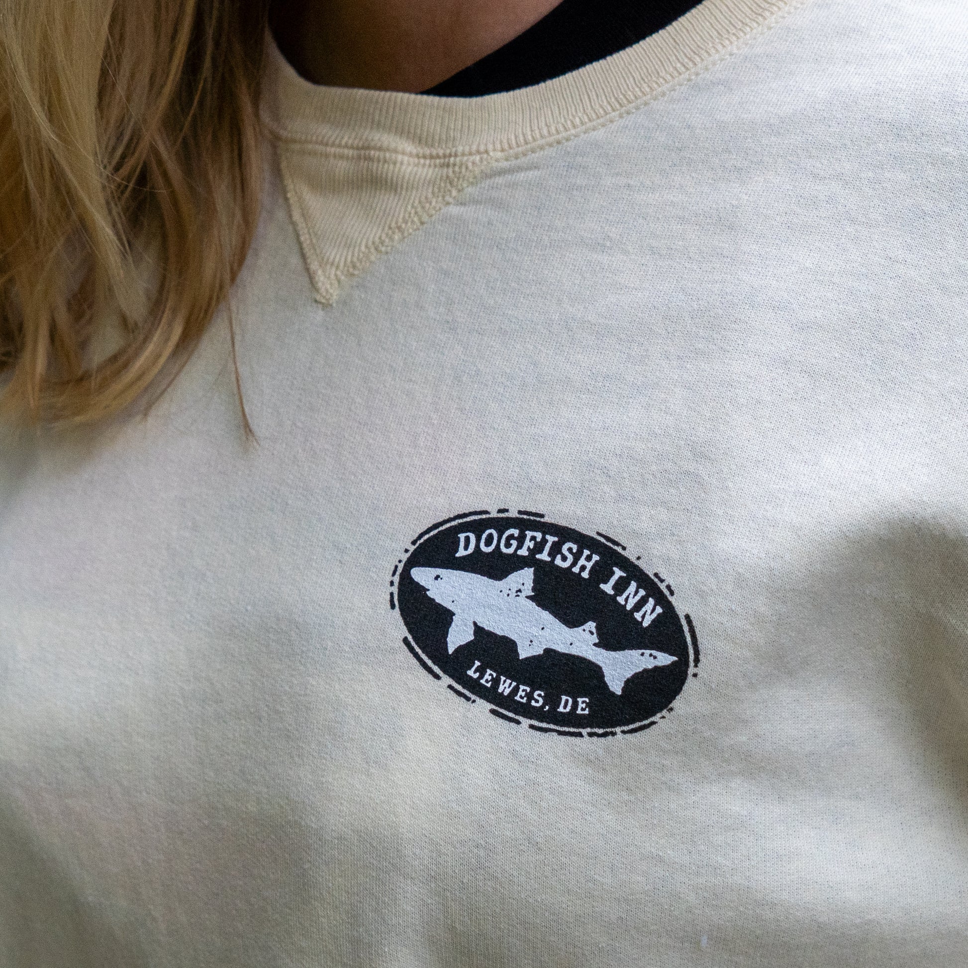 Close-up of a person wearing the Inn Cream Compass Crew by GO WEST T-SHIRT COMPANY, featuring a beer compass logo with a fish and "Dogfish Inn Lewes, DE" on the left chest. Part of their long blonde hair is visible.