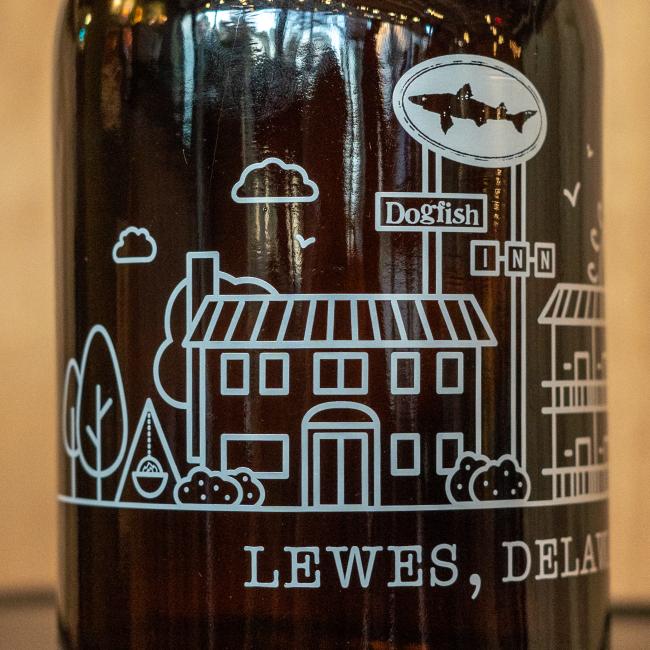 The Inn Growler 32oz by Grandstand Glassware & Apparel is a brown growler with a white line drawing of a house, trees, clouds, the Dogfish Head INN sign with fish logo, and "LEWES, DELAWARE" at the bottom.
