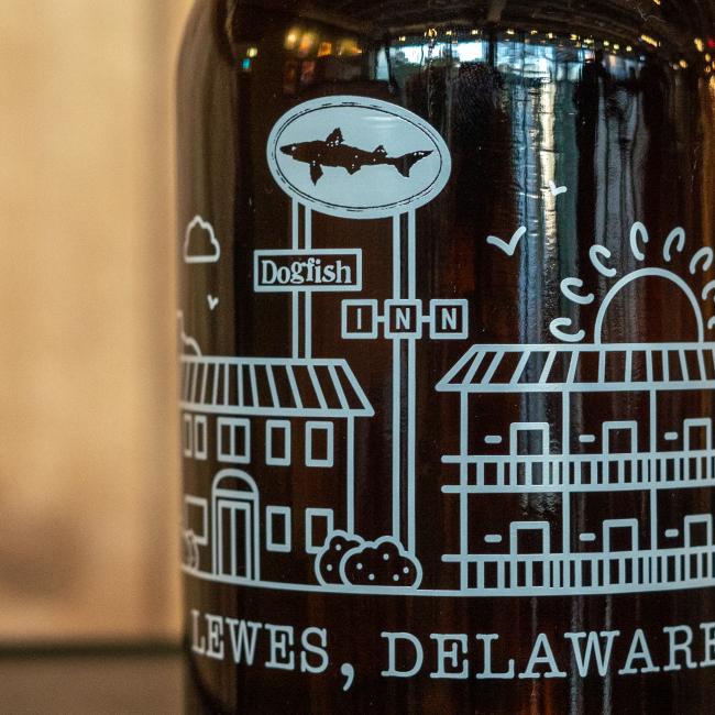A close-up of the Inn Growler 32oz by Grandstand Glassware & Apparel, featuring a white illustration of buildings, a sunrise, and a Dogfish Head INN sign above LEWES, DELAWARE.