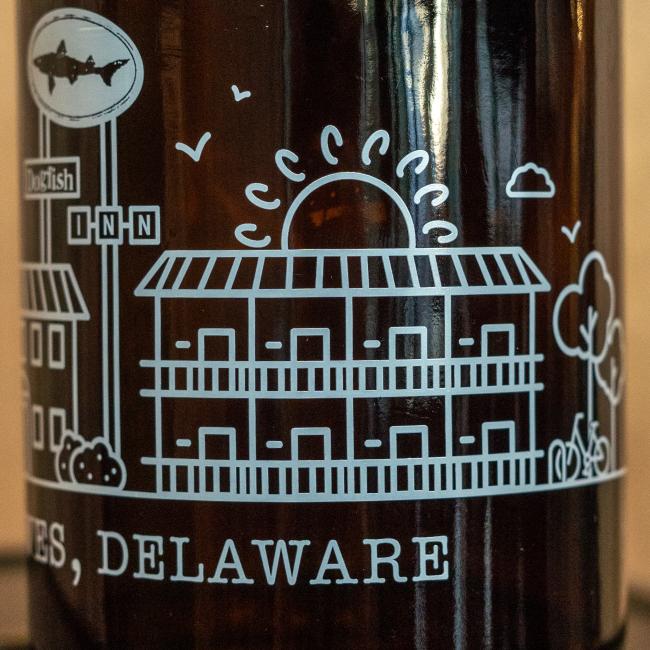 A line drawing on brown features a motel building, rising sun, birds, tree, cyclist, “INN” sign with a shark—evoking the Dogfish Head INN; partial “...ES, DELAWARE” appears below. Product: Inn Growler 32oz by Grandstand Glassware & Apparel.