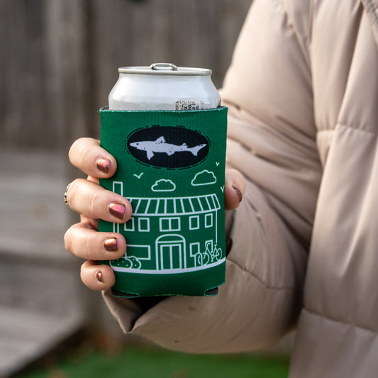 A person wearing a beige jacket holds a canned drink in the Inn Logo Can Cooler by Grandstand Glassware & Apparel, featuring a house, bicycles, clouds, and a shark silhouette—ideal for fans of unique beer accessories.