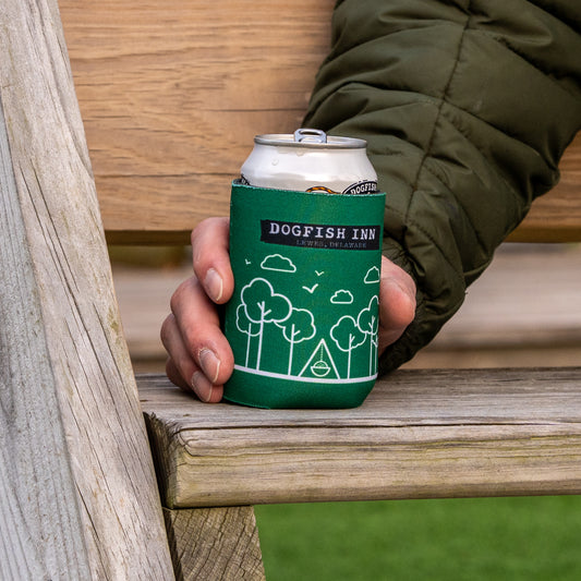 Someone in a green jacket holds a canned drink inside a Grandstand Glassware & Apparel Inn Logo Can Cooler with white tree and cloud designs, while sitting on a wooden bench.