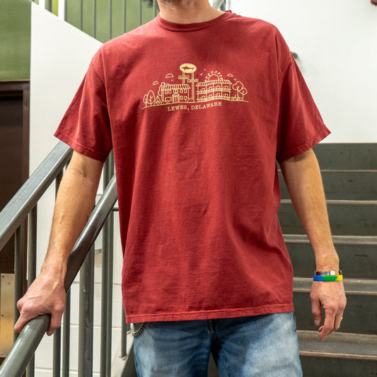 A person stands on a staircase in blue jeans and the GO WEST T-SHIRT COMPANY Inn Red Line Art Tee, featuring a simple Lewes, Delaware line art design. A colorful bracelet adds craft flair to their look. Their face is not visible.